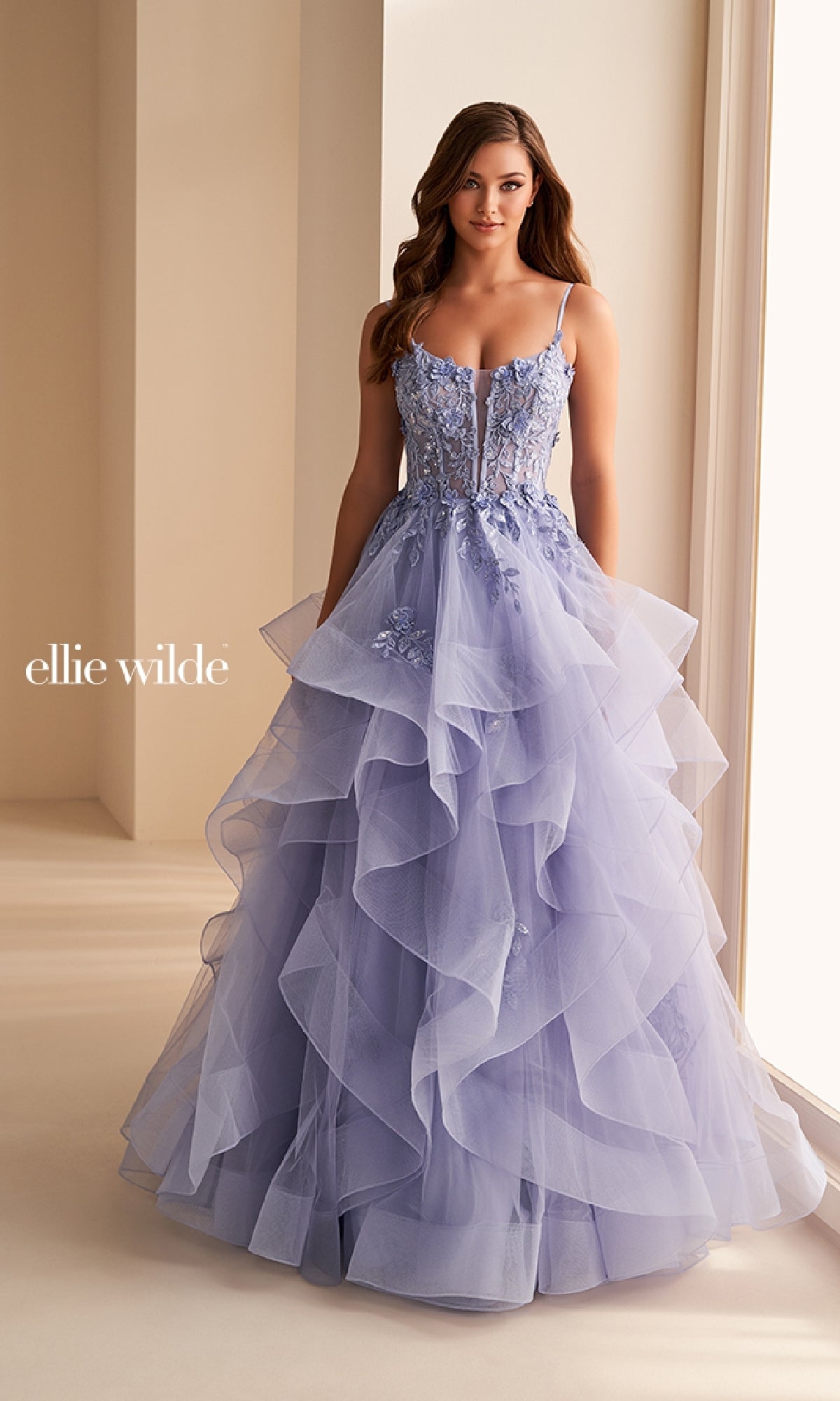 A woman in the Ellie Wilde EW36225 long prom dress featuring lavender layers, floral lace detail, and spaghetti straps stands indoors by a large window.